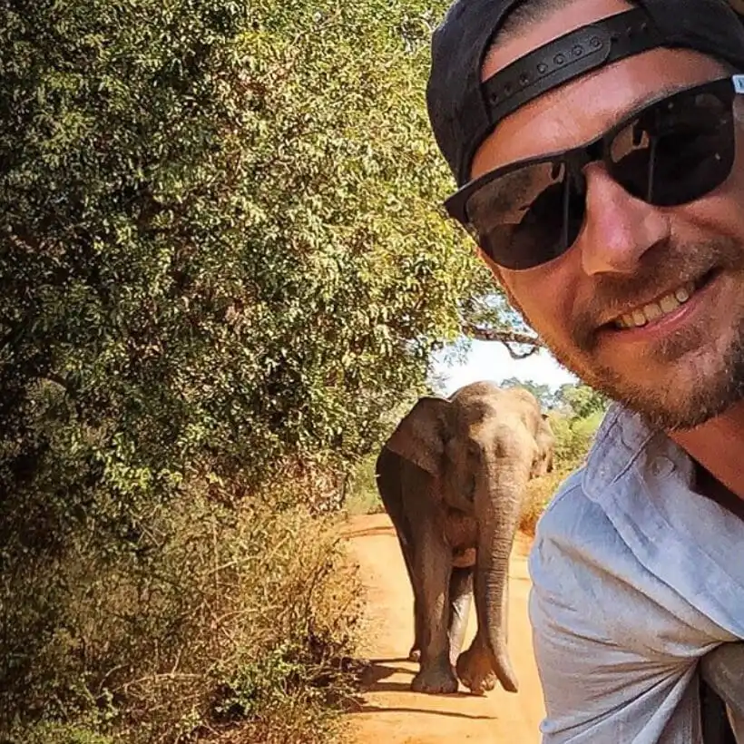 Sri Lanka Travel Guide - Ein lächelnder Mann mit Sonnenbrille und Schirmmütze macht ein Selfie auf einem unbefestigten Weg, während hinter ihm ein großer Elefant zwischen Bäumen und Büschen spaziert. - Travel Eat Love - Sri Lanka