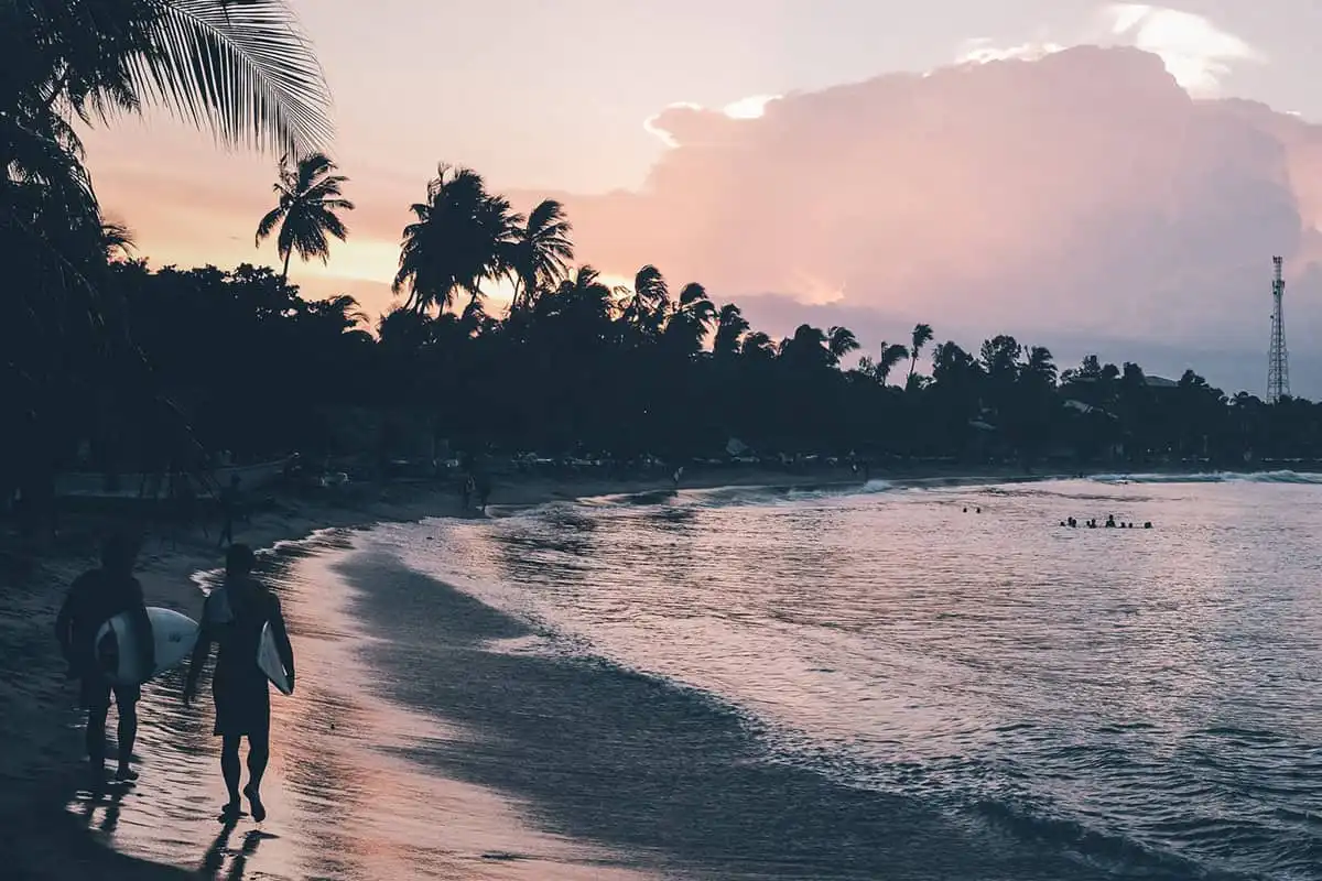 Sri Lanka Travel Guide - Zwei Personen mit Surfbrettern spazieren bei Sonnenuntergang an einem tropischen Strand entlang, an dem sich Palmen vor einem farbenfrohen Himmel abheben und sanfte Wellen an das Ufer schlagen. In der Ferne schwimmen und entspannen sich andere Menschen im Wasser. - Travel Eat Love - Sri Lanka