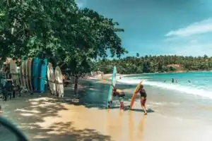 Sri Lanka Travel Guide - Am palmengesäumten tropischen Sandstrand von Strand Sri Lanka bereiten Menschen Surfbretter vor. Einige stehen oder gehen am Ufer entlang, während andere im türkisblauen Meer unter einem strahlenden, sonnigen Himmel schwimmen. - Travel Eat Love - Sri Lanka